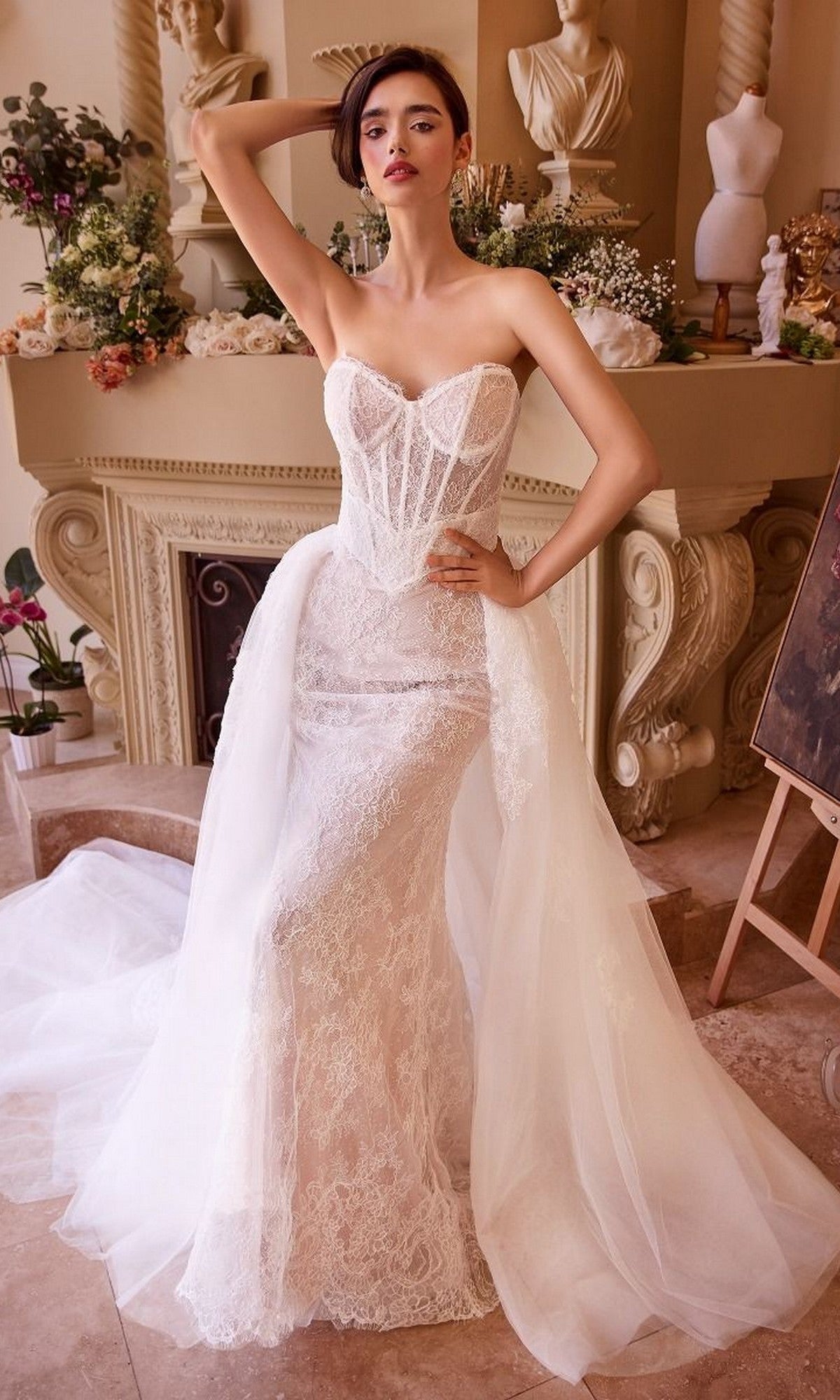 Wearing the Andrea and Leo WL044 Long Formal Dress—a strapless, lace corset gown with sheer detailing—a woman poses indoors before a decorated mantel and classical sculptures, radiating the timeless elegance of bridal celebrations.
